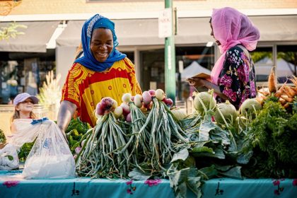 Duas mulheres conversam durante a venda de hortaliças em uma banca colorida de feira ao ar livre.