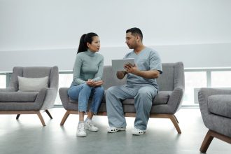 Profissional de saúde conversa com uma paciente e consulta informações em um tablet durante atendimento clínico.