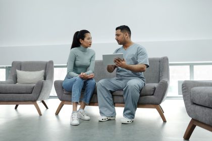 Profissional de saúde conversa com uma paciente e consulta informações em um tablet durante atendimento clínico.