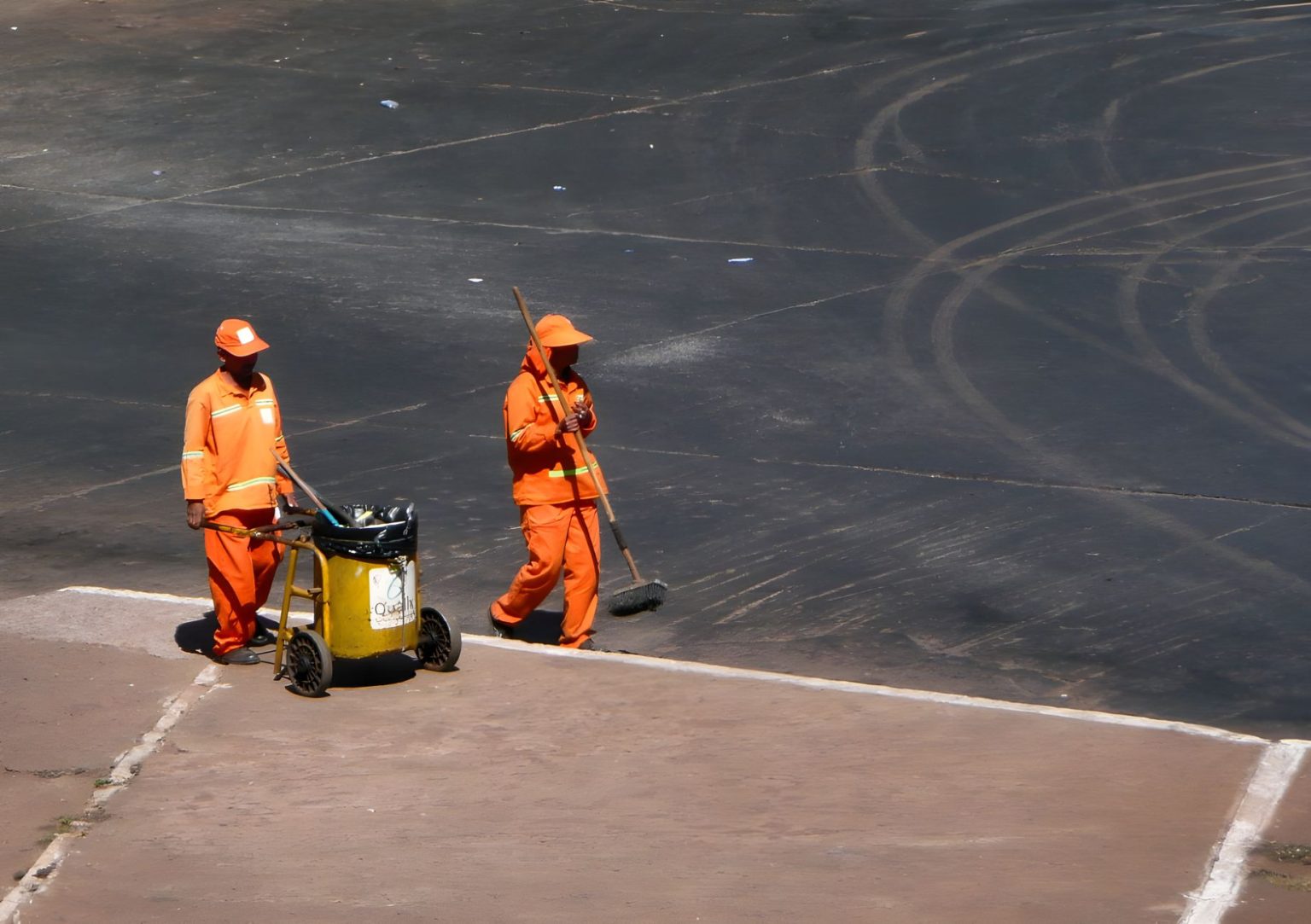 Dois trabalhadores da limpeza urbana, usando uniformes laranja, atuam em uma via da cidade.