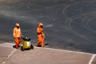 Dois trabalhadores da limpeza urbana, usando uniformes laranja, atuam em uma via da cidade.