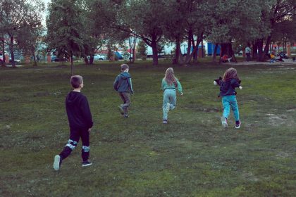 Crianças correm e brincam em uma área gramada de parque urbano cercada por árvores.